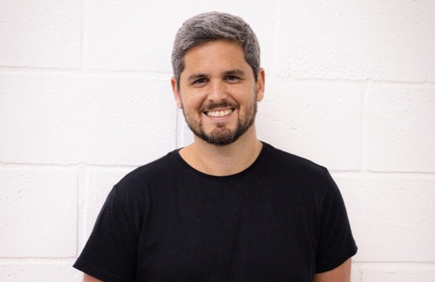 Homem sorridente de barba e cabelo grisalho, usando camiseta preta, posa contra parede de tijolos brancos.