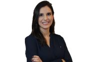 Retrato de mulher sorridente, cabelo escuro, camisa azul, braços cruzados. Fundo branco.
