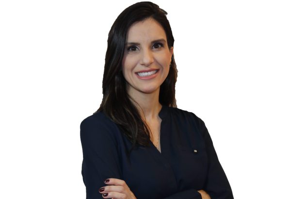 Retrato de mulher sorridente, cabelo escuro, camisa azul, braços cruzados. Fundo branco.