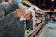 Homem branco, usando uma blusa de frio cinza e uma máscara azul, está em um supermercado lendo a nota fiscal de sua compra.