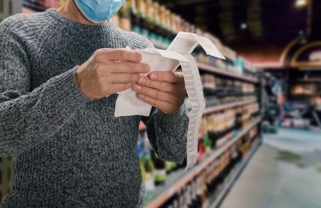 Homem branco, usando uma blusa de frio cinza e uma máscara azul, está em um supermercado lendo a nota fiscal de sua compra.