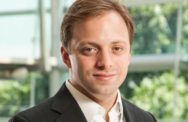 Homem de terno e camisa branca, sorrindo levemente para a câmera. Fundo corporativo.