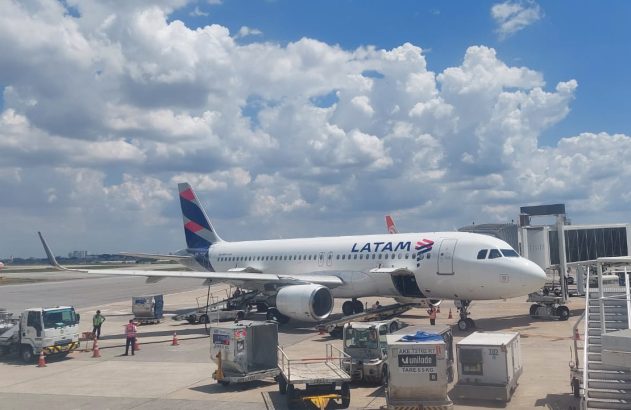 Aeronave LATAM em portão de embarque de aeroporto, com equipe de solo manuseando carga. Céu azul, dia claro.