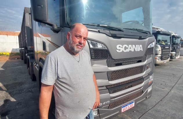 Um homem calvo e barbudo em camiseta cinza de pé ao lado de um caminhão Scania G 410 cinza, em pátio externo de dia.