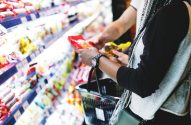 Pessoas em um supermercado, olhando e pegando produtos das prateleiras. Uma tem cabelos trançados e segura um pacote e cesta.