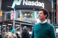 Homem sorridente em suéter verde, com o letreiro da Nasdaq visível ao fundo.