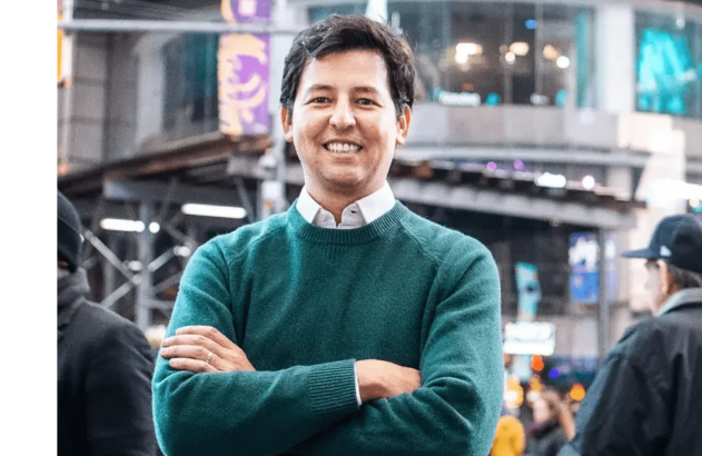 Homem de suéter verde, sorrindo e braços cruzados, em rua da cidade.