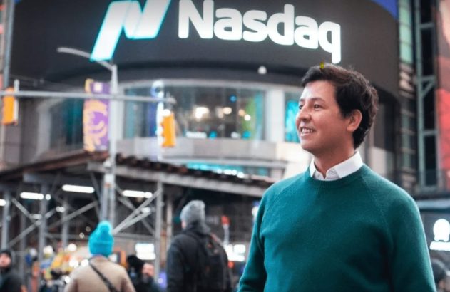 Homem sorridente em suéter verde, com o letreiro da Nasdaq visível ao fundo.