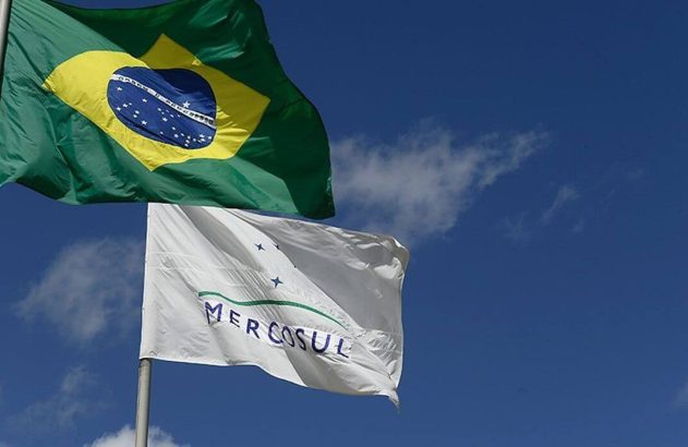 Bandeiras do Brasil e do Mercosul tremulam contra um céu azul claro com nuvens.