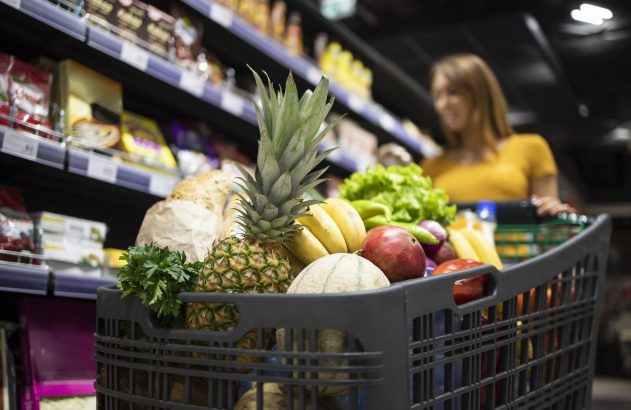 Carrinho de compras cheio de frutas, legumes e pães, sendo empurrado por mulher em corredor de supermercado.