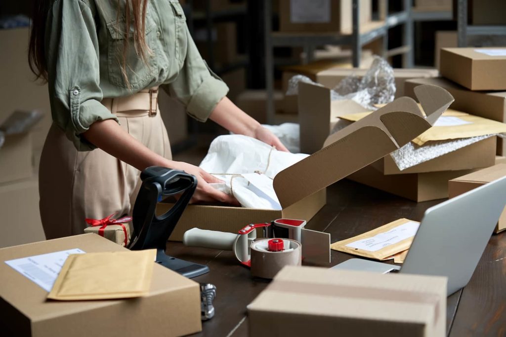A imagem mostra uma microempreendedora, vestida com uma camisa verde e uma calça bege, embalando mercadorias em uma caixa de papelão. (Foto: Shutterstock / Ground Picture)