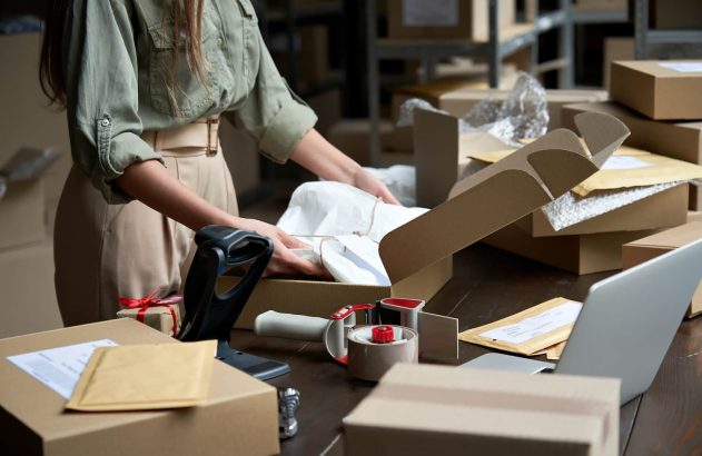 A imagem mostra uma microempreendedora, vestida com uma camisa verde e uma calça bege, embalando mercadorias em uma caixa de papelão. (Foto: Shutterstock / Ground Picture)