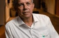 Homem grisalho de camisa branca com logo no bolso, olhando para a câmera.