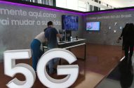 Pessoas em um estande de exposição 5G com celulares e telões informativos, e grandes letras "5G" no chão.