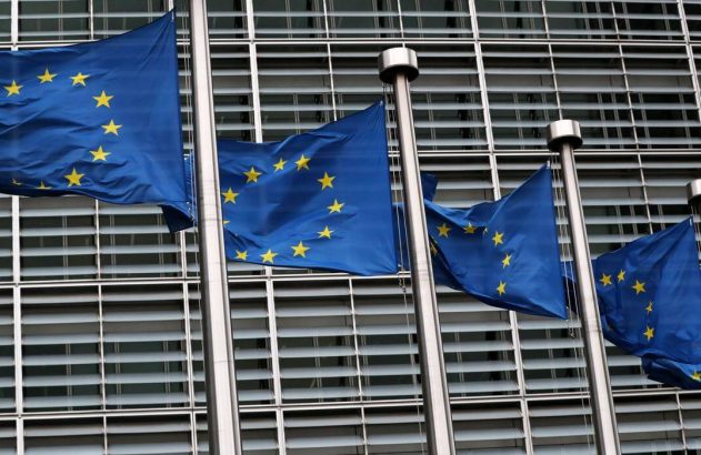 Quatro bandeiras azuis da União Europeia com estrelas amarelas tremulam em frente a um prédio moderno.