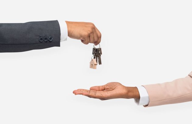 Mão de terno escuro entrega chaves com chaveiro de casa a mão de terno claro.