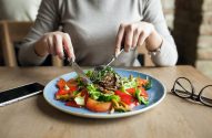Pessoa cortando salada com carne em prato azul. Há um celular e óculos na mesa.