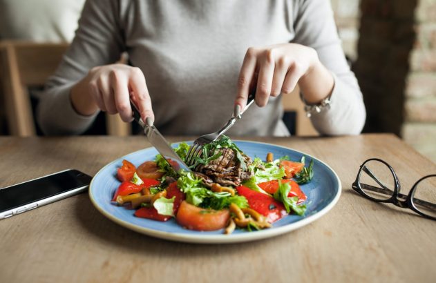 Pessoa cortando salada com carne em prato azul. Há um celular e óculos na mesa.