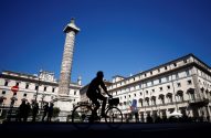 Ciclista em silhueta em Roma, com a Coluna de Marco Aurélio e o Palazzo Chigi ao fundo.