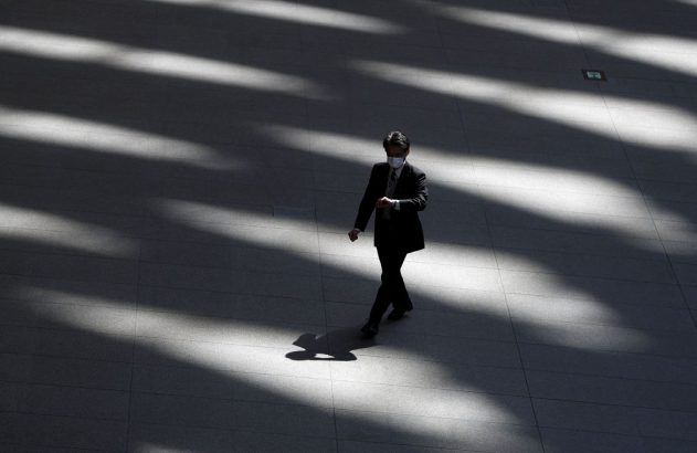 Um homem de terno e máscara caminha em piso com listras de luz e sombra. Vista de cima.
