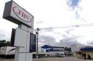 Placa "JBS" na entrada de fábrica, com edifícios industriais, caminhões e pessoas próximas ao acesso.