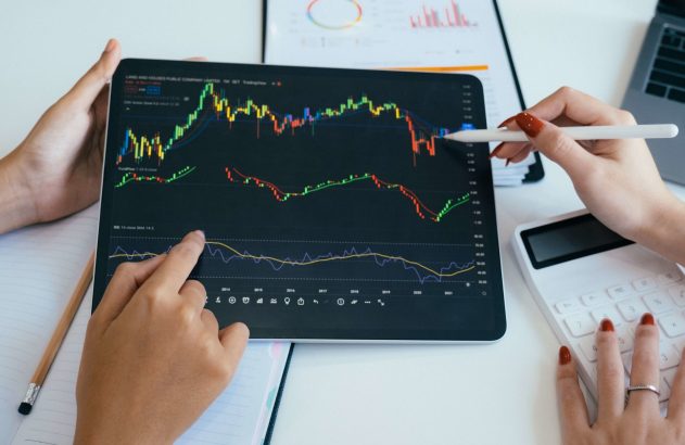 Mãos analisam gráficos financeiros em tablet com stylus, ao lado de calculadora. Análise de dados e investimentos.