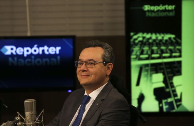 Homem de óculos e terno, sorrindo, em estúdio com microfone e telas 'Repórter Nacional'.