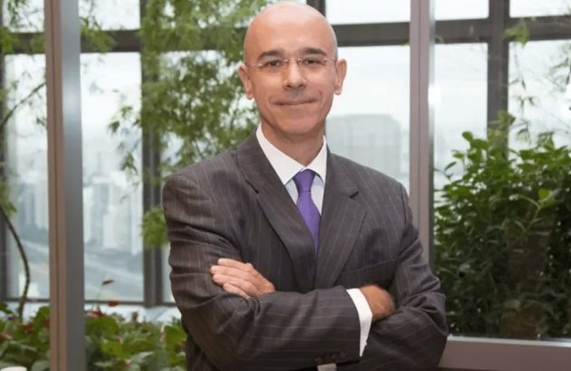 Homem de terno cinza listrado, gravata roxa, braços cruzados, sorrindo, em frente a uma janela com plantas.