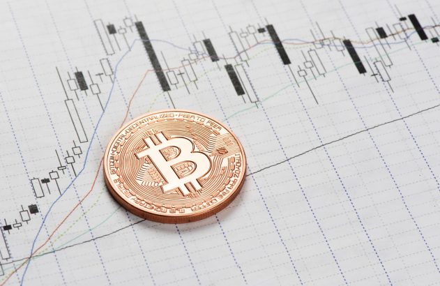Moeda dourada com o símbolo do bitcoin em cima de uma folha de papel quadriculado, em que um gráfico de linhas coloridas foi desenhado.
