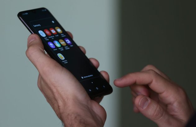 Mãos de uma pessoa segurando um smartphone Samsung. Um dedo toca a tela com aplicativos visíveis.