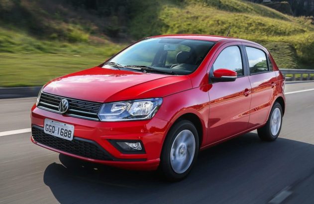 Carro hatch vermelho da Volkswagen em movimento em uma rodovia, com paisagem verde ao fundo.