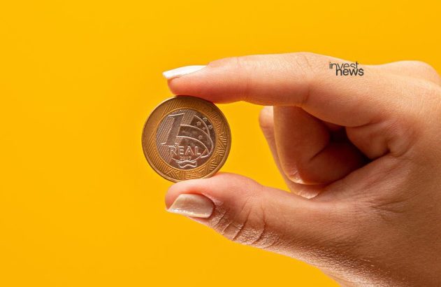 Mão segurando uma moeda de 1 Real brasileiro contra fundo amarelo.