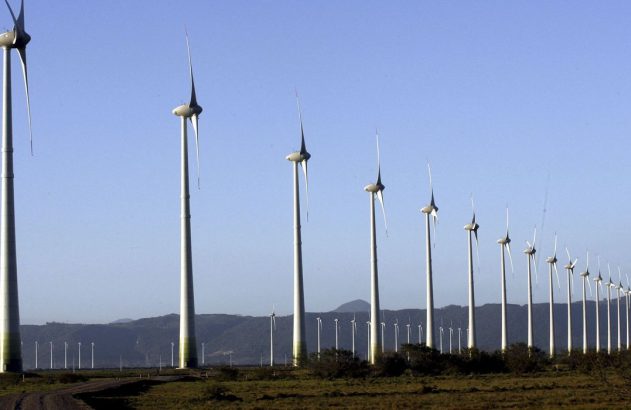Longa fileira de turbinas eólicas brancas em um campo, sob céu azul e montanhas no horizonte.