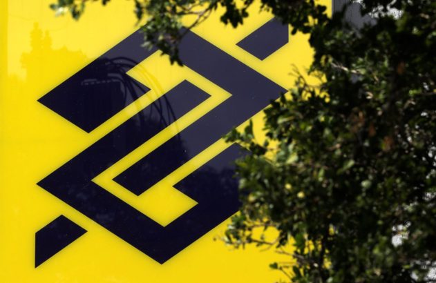 Logo azul-escuro do Banco do Brasil em fundo amarelo, com ramos de árvores cobrindo parte da imagem.