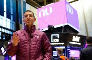 Homem em casaco roxo gesticula e fala. Ao fundo, ambiente de bolsa de valores com logos NuBank e ICE.