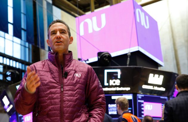 Homem em casaco roxo gesticula e fala. Ao fundo, ambiente de bolsa de valores com logos NuBank e ICE.