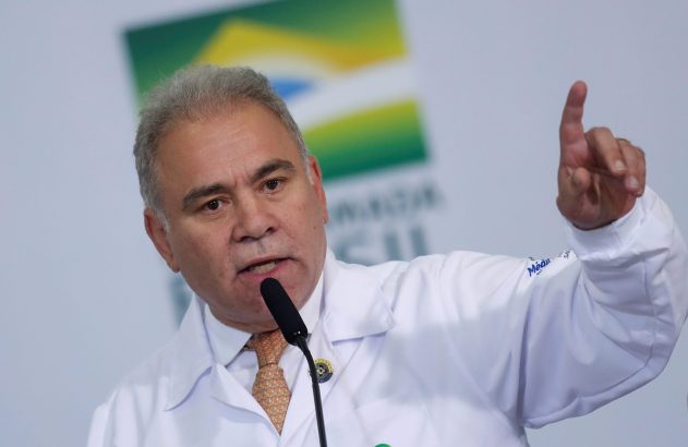 Homem de jaleco branco, provavelmente médico, fala em microfone em um evento, apontando o dedo indicador para cima.