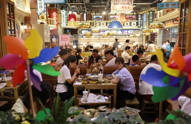 Pessoas comendo em um restaurante movimentado, com letreiros chineses e moinhos de vento coloridos no primeiro plano.