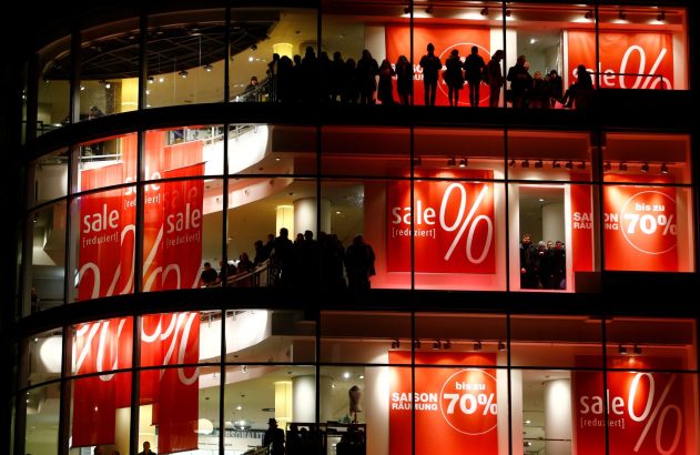 Loja iluminada à noite, com vários andares, pessoas e cartazes vermelhos de "SALE" e "até 70% de desconto".