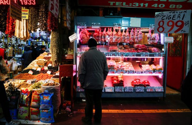 Homem em mercado vibrante, olhando balcão de carnes frescas e especiarias. Placas com preços em turco.