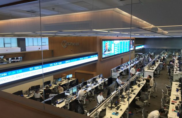 Corretora de valores BTG Pactual, com funcionários em mesas e grandes telas exibindo dados financeiros.