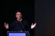 Jair Bolsonaro discursando em um púlpito com microfones, gesticulando com as mãos abertas, em um fundo escuro.