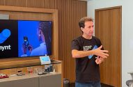 Homem em camiseta "desbloqueie seu mundo crypto" gesticula. TV da Mynt com mulher ao telefone no fundo.