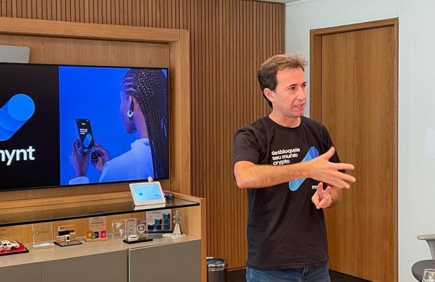 Homem em camiseta "desbloqueie seu mundo crypto" gesticula. TV da Mynt com mulher ao telefone no fundo.