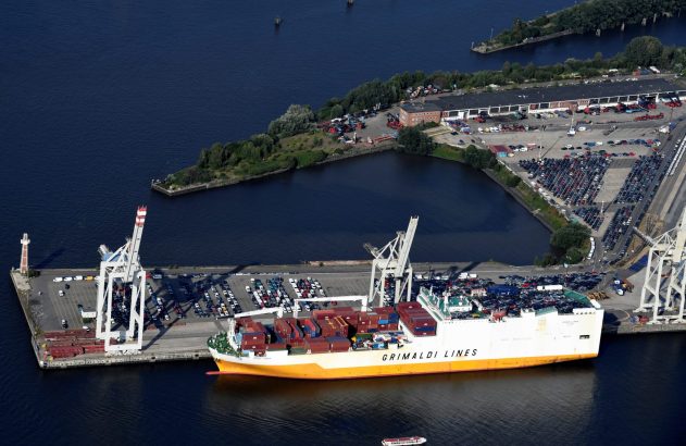 Vista aérea: navio Grimaldi Lines carregado com carros e contêineres atracado em doca com pátios de carros e guindastes.