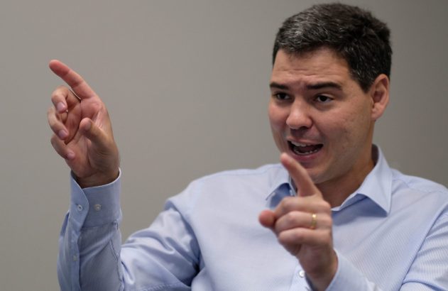 Homem de camisa azul clara gesticula com as duas mãos, apontando com os indicadores enquanto fala.