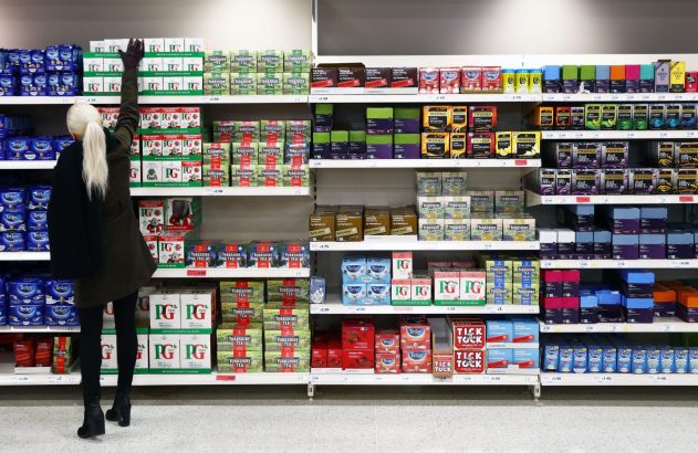 Mulher alcança caixa de chá em prateleira superior de supermercado.