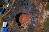 Vista aérea de garimpo: grande cratera em terra vermelha, áreas desmatadas e queimadas, mangueiras, acampamento com tendas.
