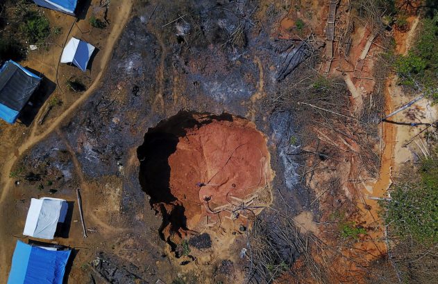 Vista aérea de garimpo: grande cratera em terra vermelha, áreas desmatadas e queimadas, mangueiras, acampamento com tendas.