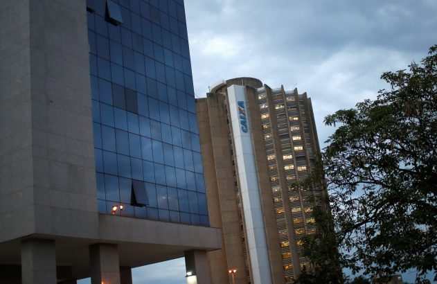 Dois prédios sob céu nublado e árvore à direita. Um com janelas de vidro abertas, outro com 'CAIXA' e luzes.